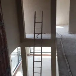 Surowe wnętrze dwupoziomowego domu w trakcie prac wykończeniowych; widoczna drewniana drabina oparta o otwór w stropie, ściany pokryte świeżym tynkiem, kable elektryczne ułożone wzdłuż podłogi...