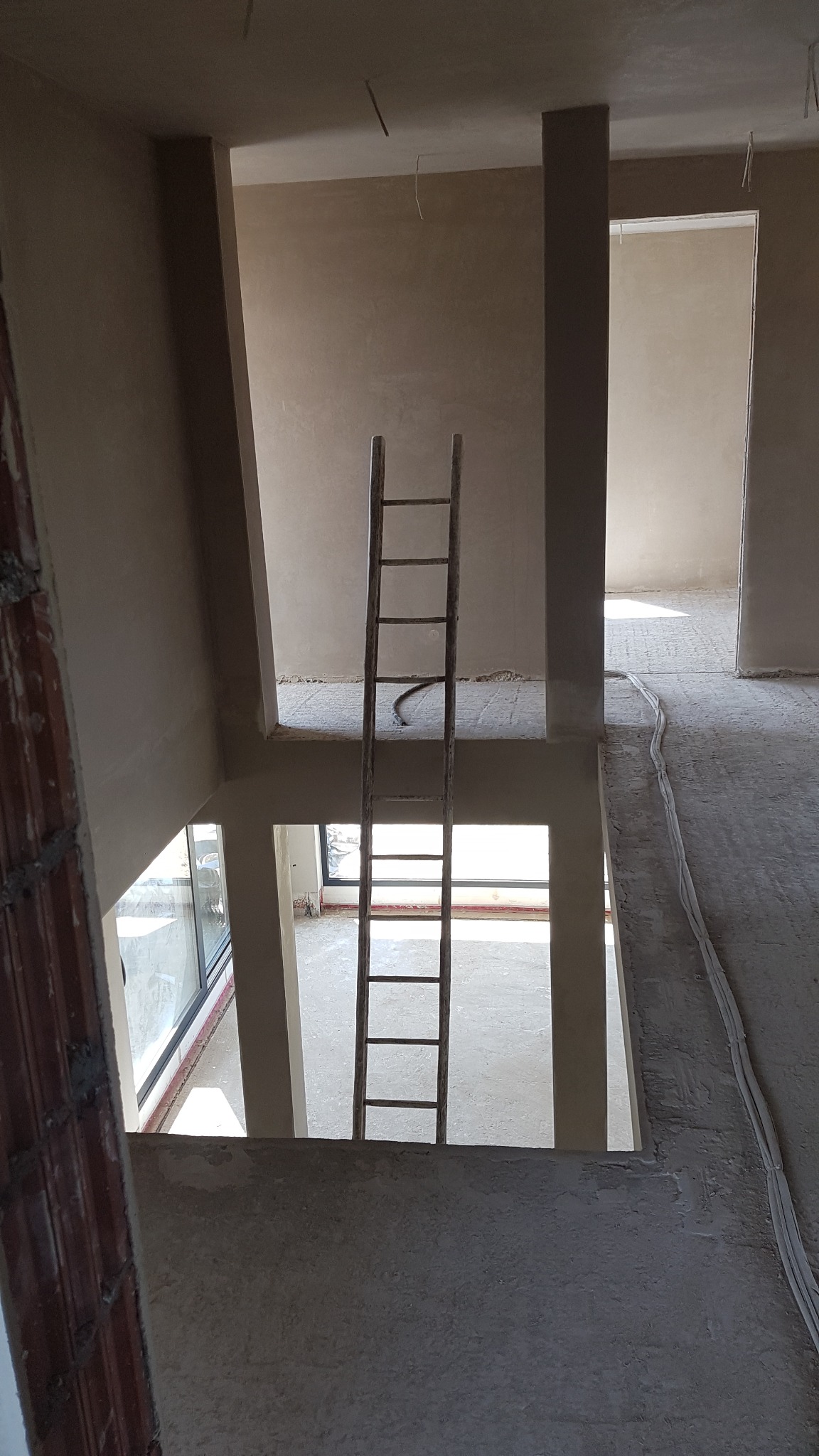 Surowe wnętrze dwupoziomowego domu w trakcie prac wykończeniowych; widoczna drewniana drabina oparta o otwór w stropie, ściany pokryte świeżym tynkiem, kable elektryczne ułożone wzdłuż podłogi...