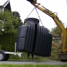 Żółty minikoparka Zeppelin podnosi duży, czarny, żebrowany zbiornik z żółtą naklejką, zawieszony na linach, obok zaparkowana przyczepka, w tle dom i zieleń.