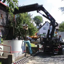 Montaż betonowego zbiornika za pomocą dźwigu samochodowego na posesji z domem w tle, widoczni pracownicy w strojach roboczych.