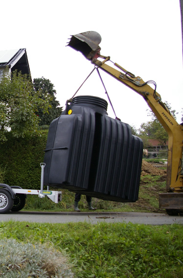Żółty minikoparka Zeppelin podnosi duży, czarny, żebrowany zbiornik z żółtą naklejką, zawieszony na linach, obok zaparkowana przyczepka, w tle dom i zieleń.