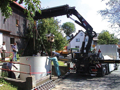 Montaż betonowego zbiornika za pomocą dźwigu samochodowego na posesji z domem w tle, widoczni pracownicy w strojach roboczych.