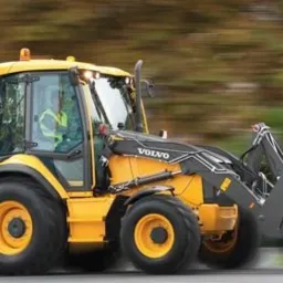 Żółto-czarna koparko-ładowarka Volvo BL71B w ruchu, z rozmytym tłem drzew, widoczna z boku, operator w kabinie, dynamiczne ujęcie.