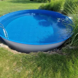 Okrągły, naziemny basen z niebieską mozaiką wewnątrz, częściowo zagłębiony w ziemi na trawniku, z widocznymi kablami i rurami przy krawędzi wykopu.