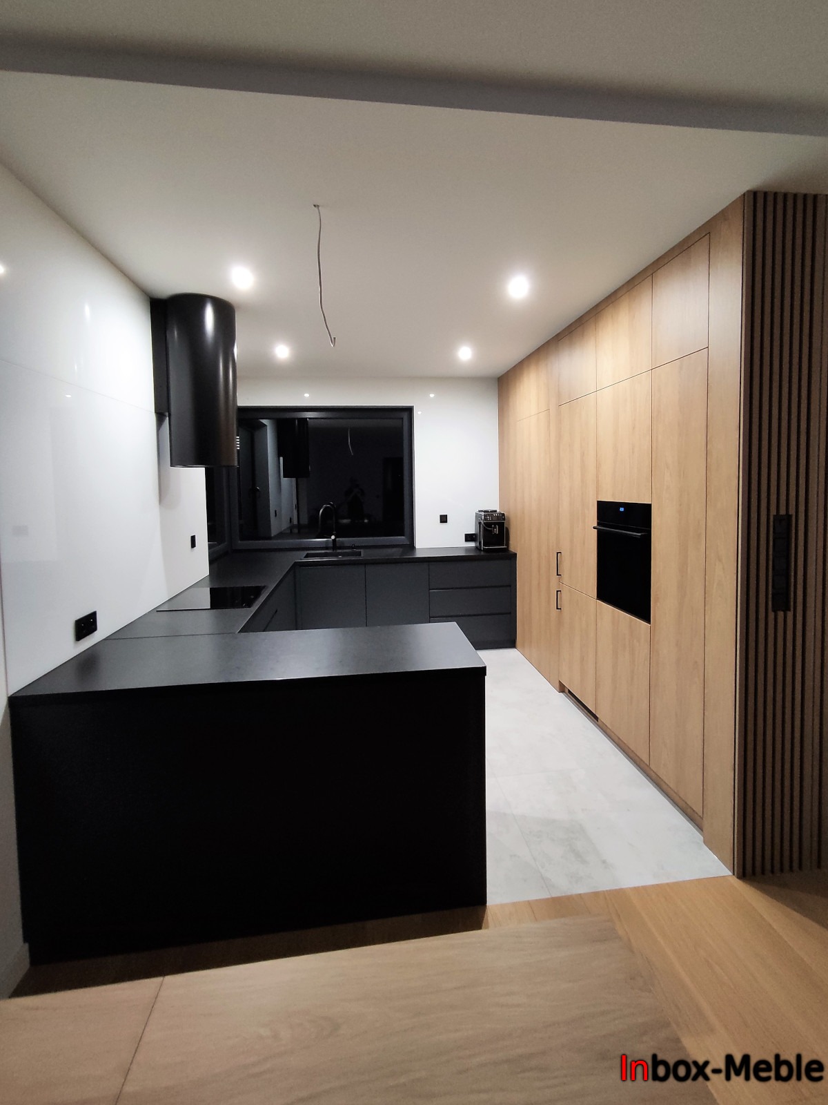 Nowoczesna kuchnia z czarnymi blatami i frontami oraz drewnianą zabudową ścienną, widok na wyspę kuchenną i okno, minimalistyczny design.