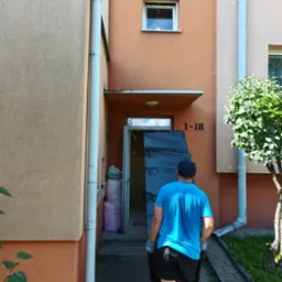 Mężczyzna w niebieskiej koszulce i rękawicach wnosi mebel owinięty folią do budynku z numerem 1-18.