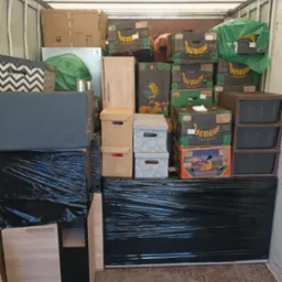 Wnętrze busa wypełnione po brzegi kartonami różnych rozmiarów, meblami owiniętymi czarną folią stretch i workami, gotowe do transportu podczas przeprowadzki.