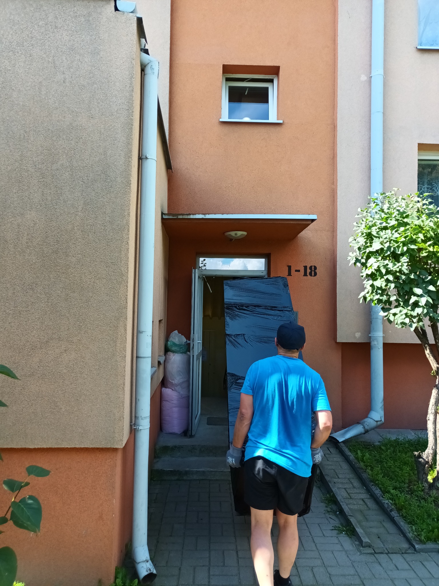 Mężczyzna w niebieskiej koszulce i rękawicach wnosi mebel owinięty folią do budynku z numerem 1-18.