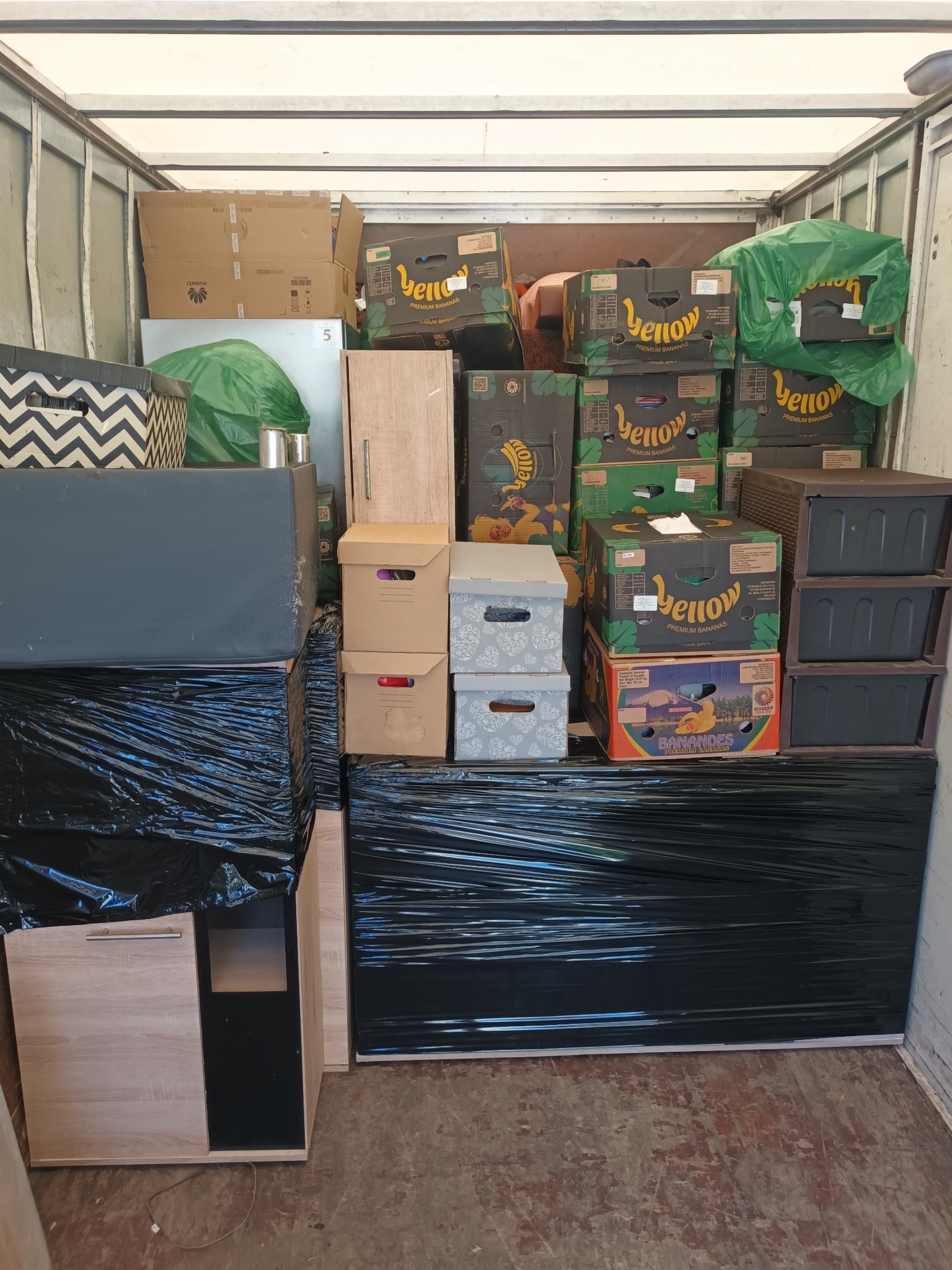 Wnętrze busa wypełnione po brzegi kartonami różnych rozmiarów, meblami owiniętymi czarną folią stretch i workami, gotowe do transportu podczas przeprowadzki.