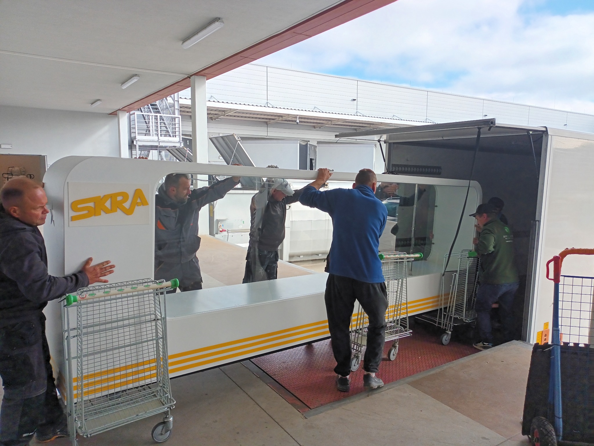 Ekipa w Łasku ładuje wózki transportowe do przyczepy z logo SKRA, widoczny fragment rampy załadunkowej i pracownicy w trakcie załadunku.
