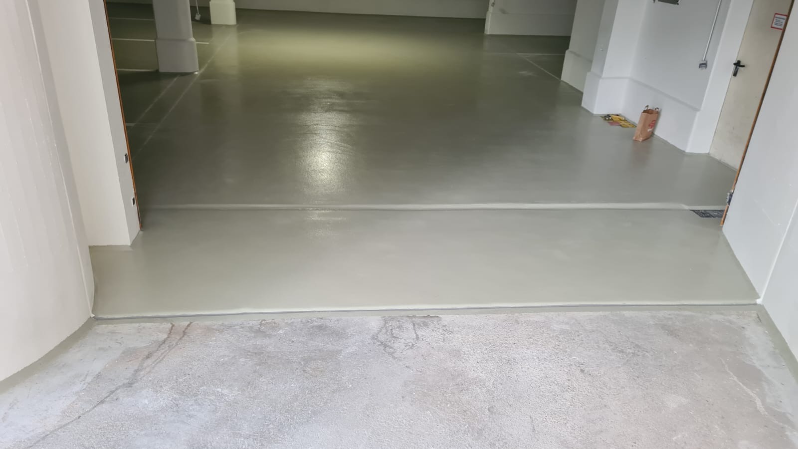 Jasnoszara, gładka podłoga w hali magazynowej, widoczny próg oddzielający starą i nową powierzchnię, w tle torba papierowa i narzędzia.