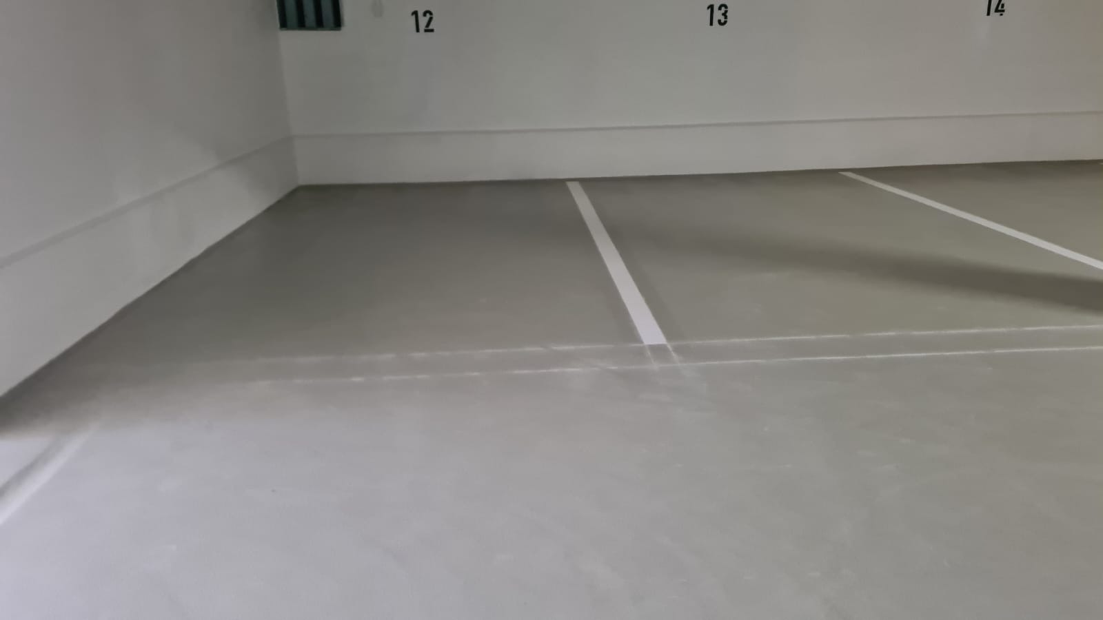 Szara, gładka powierzchnia podłogi w garażu z namalowanymi białymi liniami wyznaczającymi miejsca parkingowe, numery miejsc 12, 13 i 14 widoczne na ścianie.