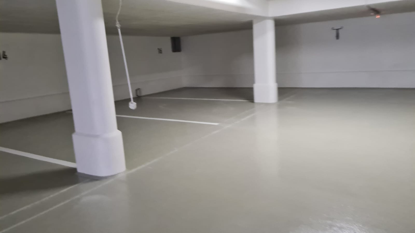 Jasnoszara, błyszcząca podłoga w garażu podziemnym z białymi liniami wyznaczającymi miejsca parkingowe, wsparta na białych, kwadratowych filarach.