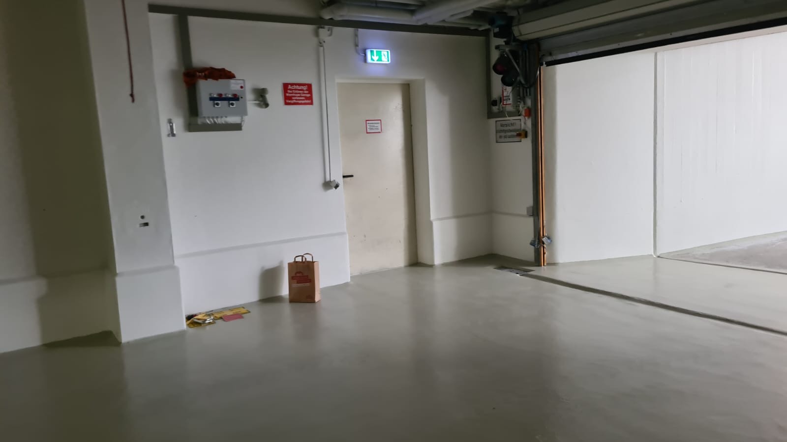 Szara, gładka posadzka w garażu z otwartą bramą, przy ścianie torba papierowa i ulotki, na ścianie tablica ostrzegawcza i skrzynka elektryczna.