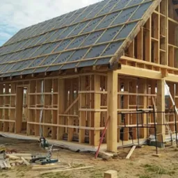 Konstrukcja drewniana domu w trakcie budowy z widocznymi elementami więźby dachowej pokrytej membraną, na pierwszym planie narzędzia budowlane i resztki drewna.