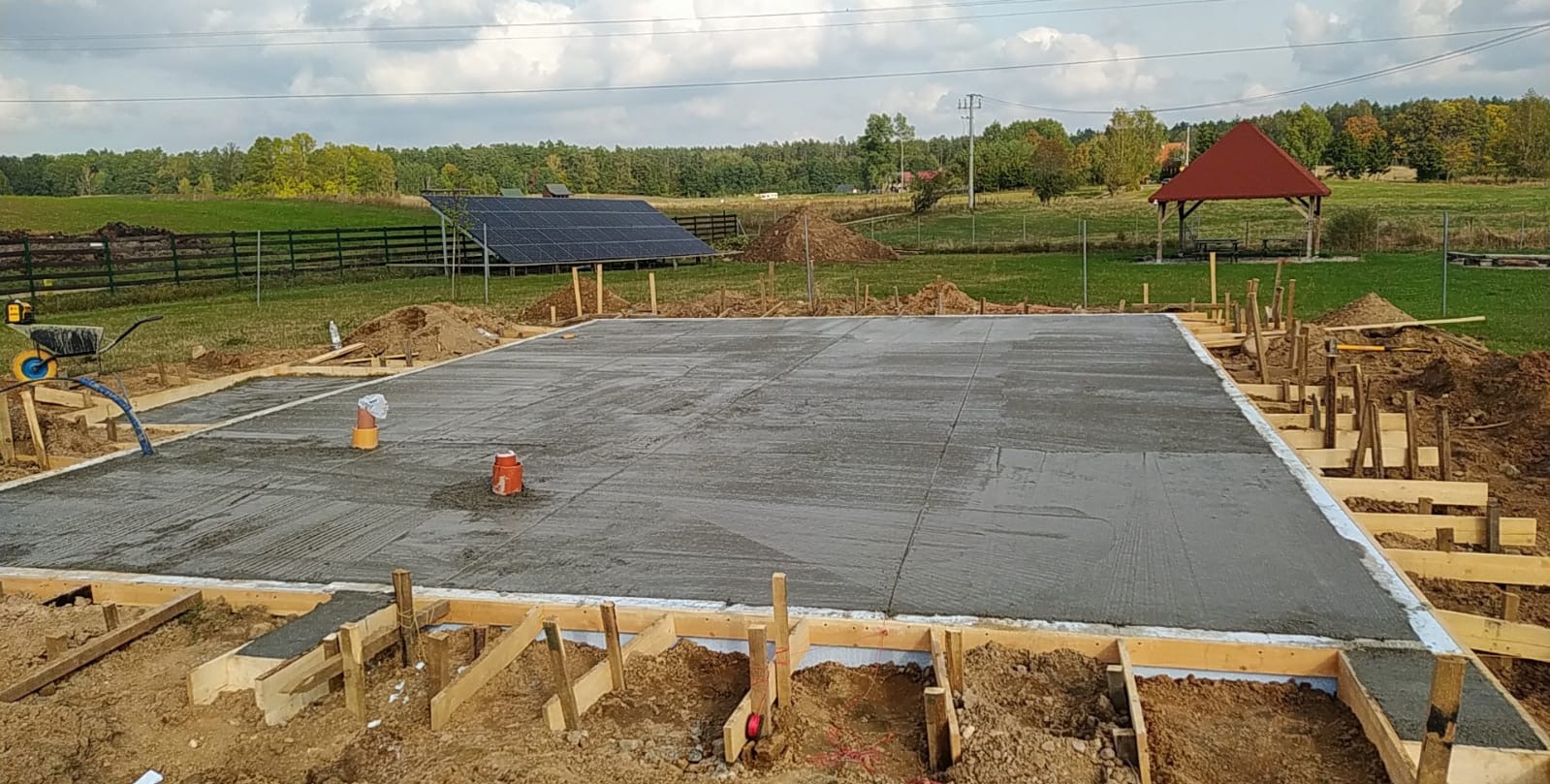 Wylana płyta fundamentowa pod budynek, z widocznym szalunkiem, izolacją termiczną i wystającymi rurami kanalizacyjnymi na tle zielonego krajobrazu z panelami słonecznymi i altaną.