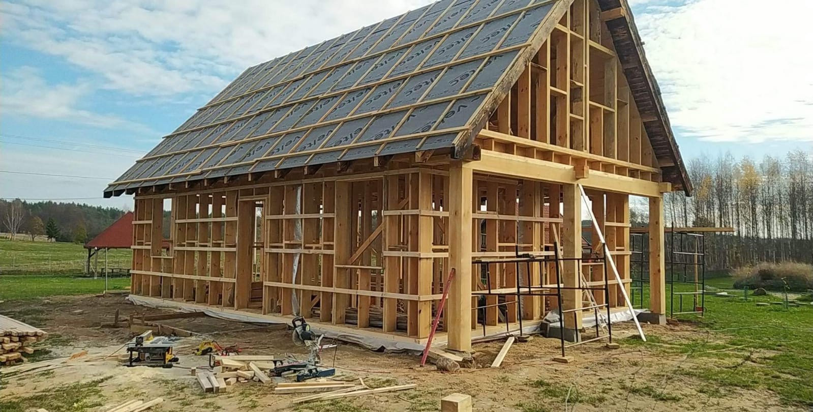 Konstrukcja drewniana domu w trakcie budowy z widocznymi elementami więźby dachowej pokrytej membraną, na pierwszym planie narzędzia budowlane i resztki drewna.