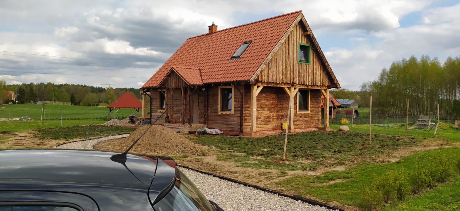 Surowy, drewniany dom z czerwoną dachówką w wiejskim otoczeniu, widok zza samochodu, świeżo posiana trawa w ogrodzie.