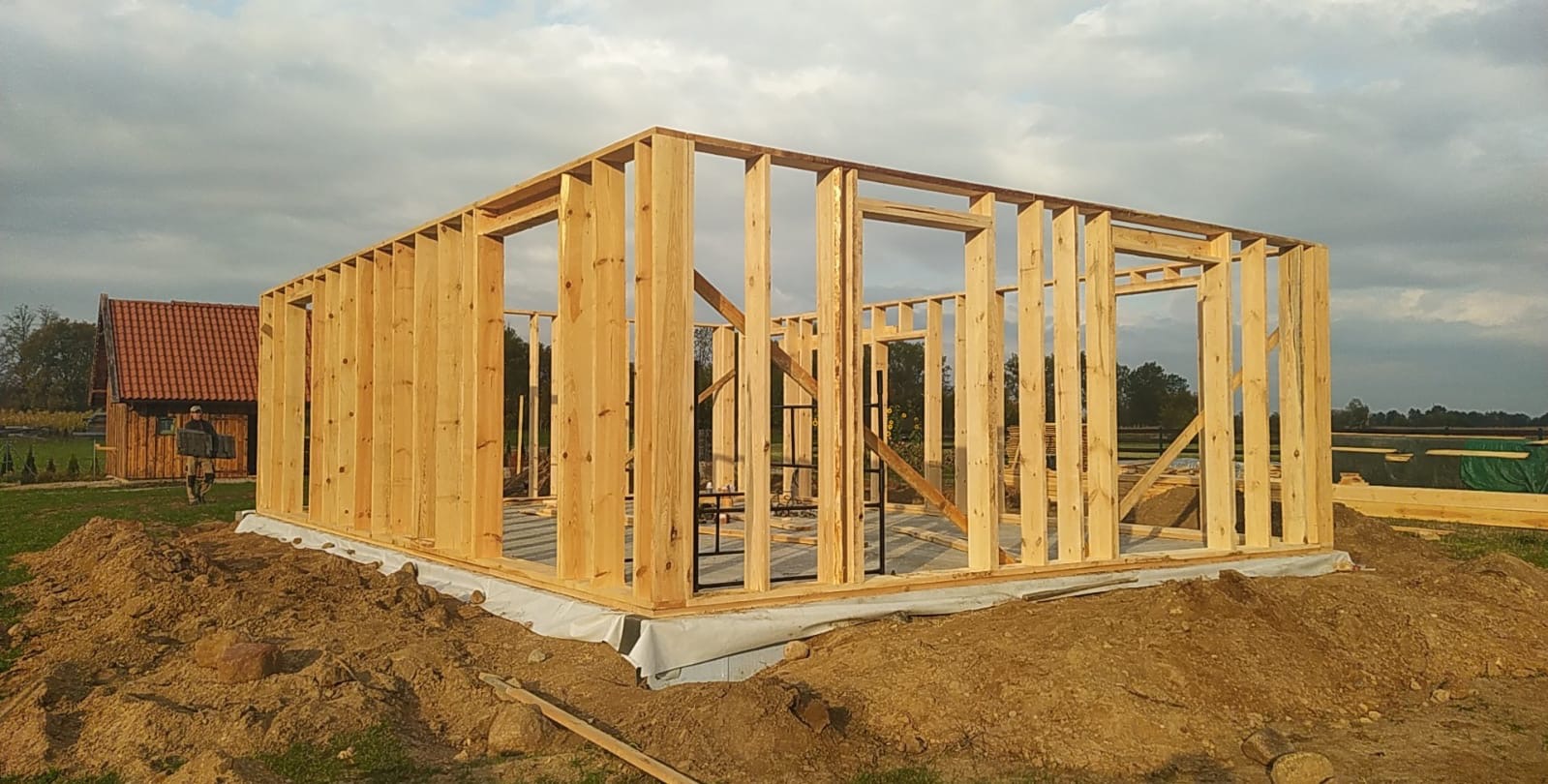 Konstrukcja szkieletowa domu w trakcie budowy, widoczne drewniane ramy ścian i stropu na fundamencie z hydroizolacją, w tle budynek z czerwonym dachem i postać przenosząca materiały budowlane.