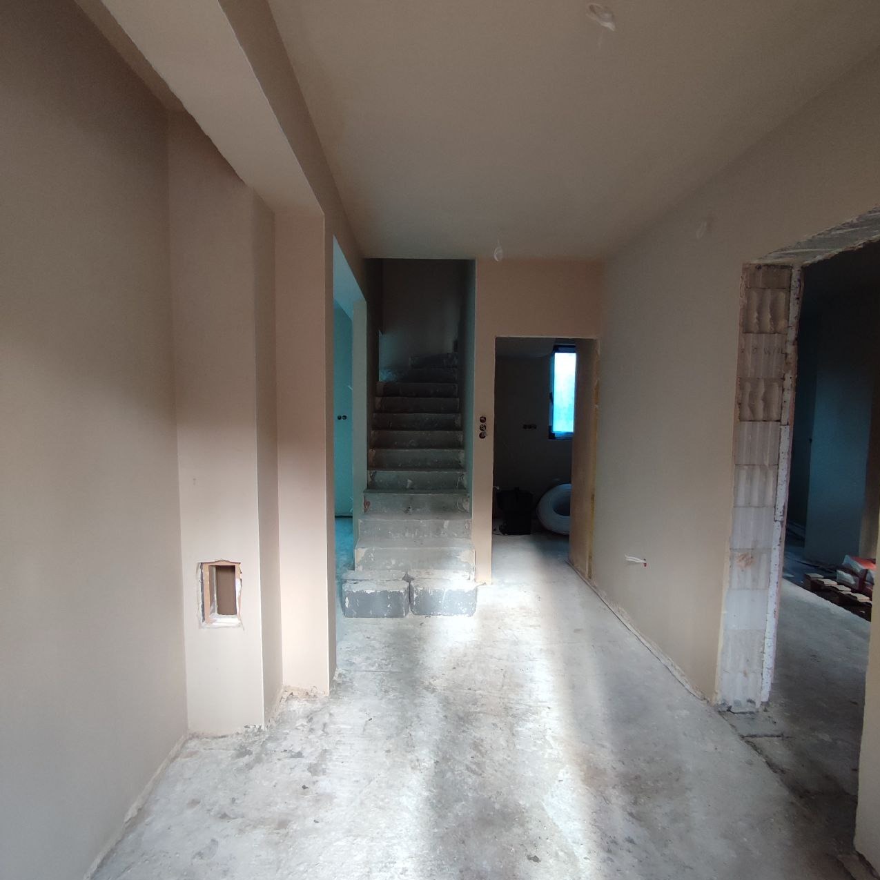 Wnętrze w trakcie prac wykończeniowych: świeżo otynkowane ściany w odcieniach beżu, surowy beton na podłodze, widoczne schody z szarego betonu prowadzące na wyższą kondygnację,...