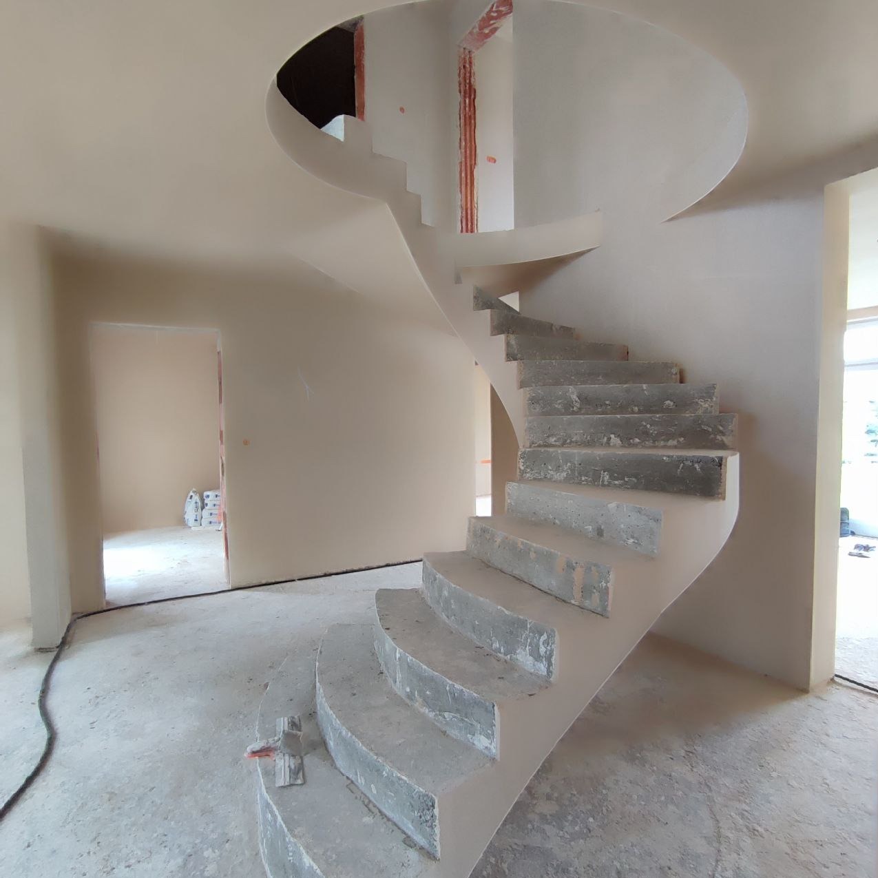 Wnętrze w trakcie prac wykończeniowych: spiralne schody z surowego betonu prowadzą na wyższą kondygnację, ściany pokryte świeżym, jasnym tynkiem, widoczne worki z materiałami budowlanymi w oddali.
