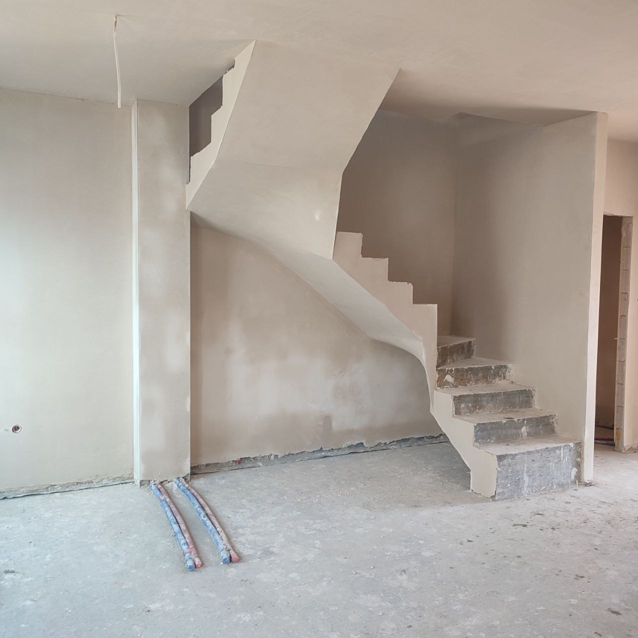Wnętrze w trakcie prac wykończeniowych: schody i ściany pokryte świeżym, jasnym tynkiem, widoczne przewody na podłodze.
