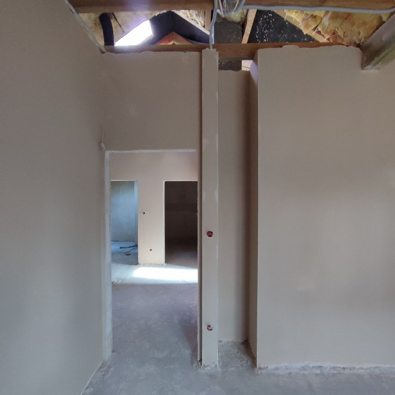 Wnętrze w trakcie prac wykończeniowych: świeżo nałożone, jasne tynki na ścianach, widoczne otwory drzwiowe, surowy strop z instalacjami. Na ścianie widoczne punkty instalacji elektrycznej.