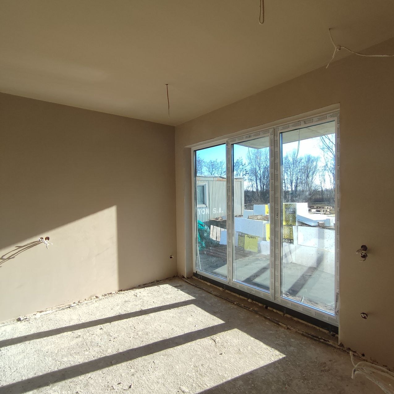 Świeżo otynkowane wnętrze z dużym oknem przesuwnym, widoczne kable elektryczne wystające ze ściany, podłoga w stanie surowym, na zewnątrz budynku widoczne materiały budowlane i kontener biurowy.