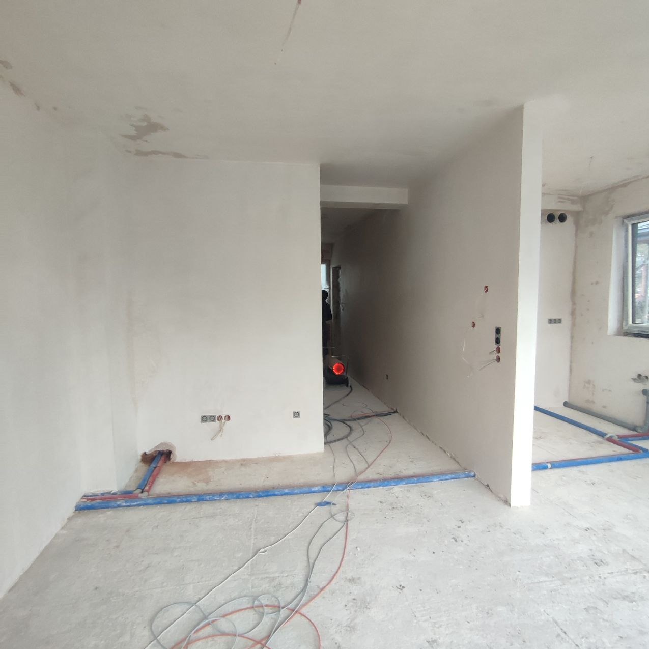 Wnętrze w trakcie prac tynkarskich maszynowych; ściany pokryte świeżym, jasnym tynkiem, widoczne instalacje elektryczne i hydrauliczne, przewody na podłodze, w głębi pomieszczenia czerwony...