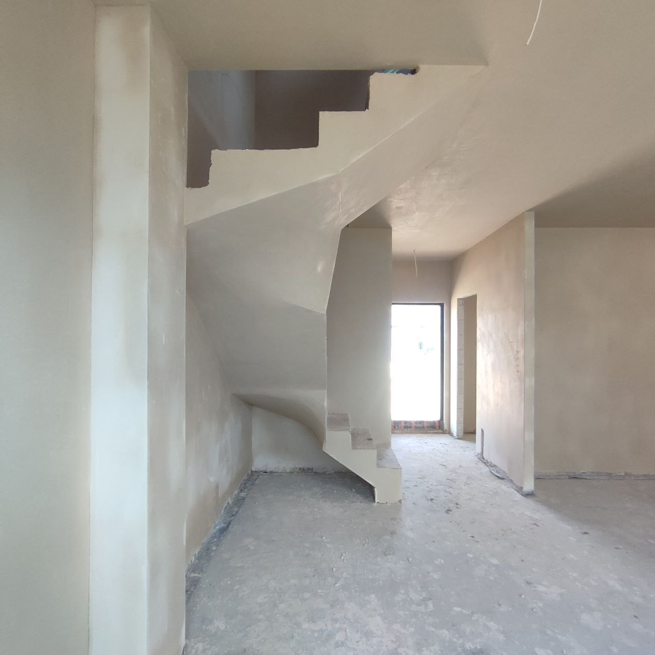Wnętrze domu w trakcie prac wykończeniowych, widoczne świeżo nałożone, jasne tynki gipsowe na ścianach i schodach, surowy beton na podłodze, oraz okno w oddali wpuszczające naturalne światło.