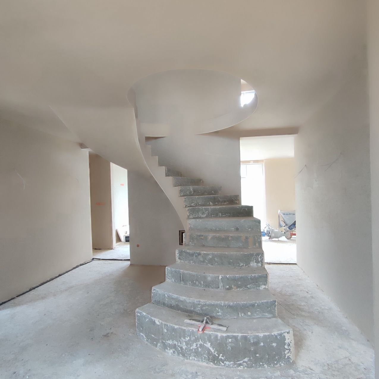 Wnętrze w trakcie prac wykończeniowych: spiralne schody z surowego betonu prowadzą na piętro, ściany pokryte świeżym, jasnym tynkiem, widoczne narzędzia murarskie na stopniach.