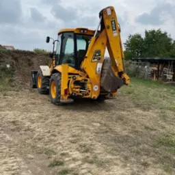 Żółta koparko-ładowarka JCB 4CX z podniesioną łyżką na nierównym, piaszczystym terenie z widoczną stertą ziemi w tle.