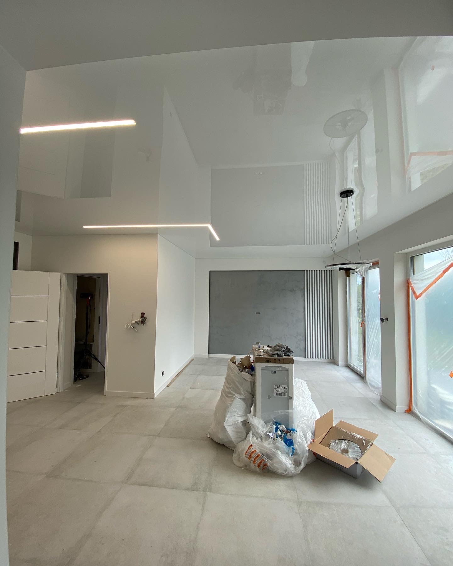 Błyszczący sufit z podłużnymi lampami LED w nowym, jasnym wnętrzu z dużymi oknami, widoczne ślady prac wykończeniowych i materiały budowlane