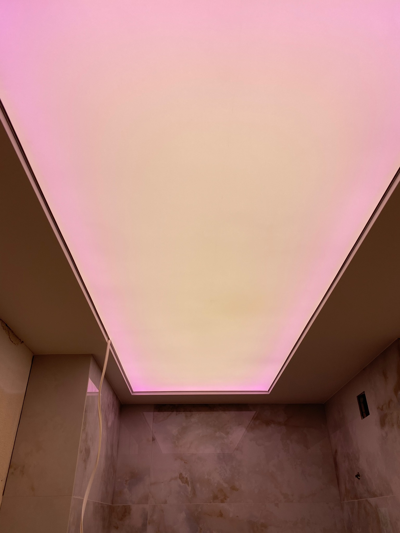 Prostokątny sufit napinany z subtelnym, różowo-żółtym podświetleniem LED, widoczny od dołu, z fragmentem ściany w odcieniach beżu i brązu.