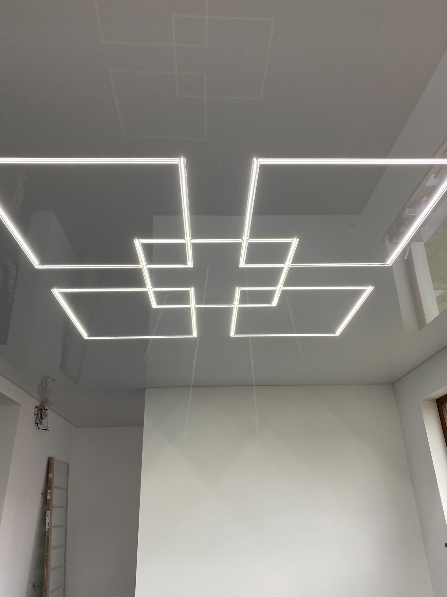 Błyszczący sufit napinany z geometrycznym wzorem wykonanym z podświetlanych profili LED, odbicie w suficie, białe ściany w tle.