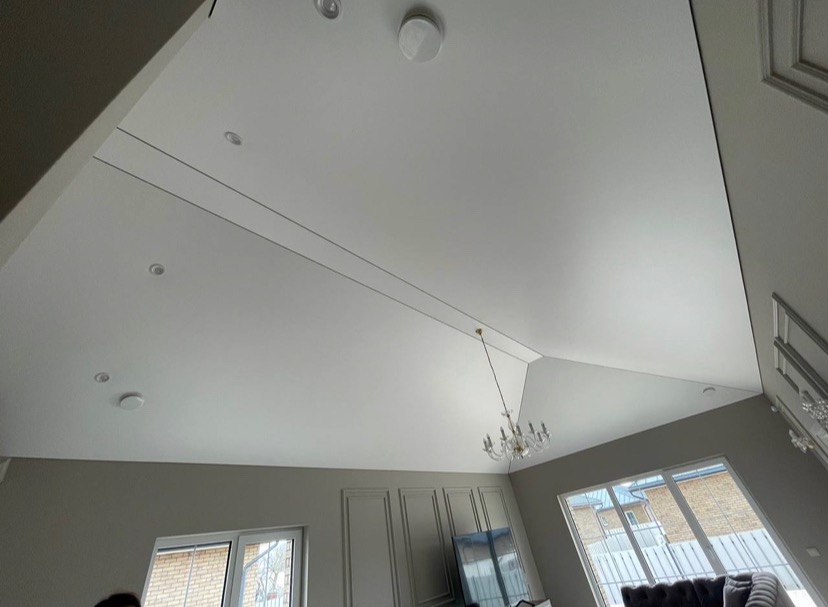 Nowoczesny, biały sufit napinany z wbudowanymi punktowymi źródłami światła i ukrytymi liniami podziału, oświetlony wiszącym żyrandolem w przestronnym salonie.