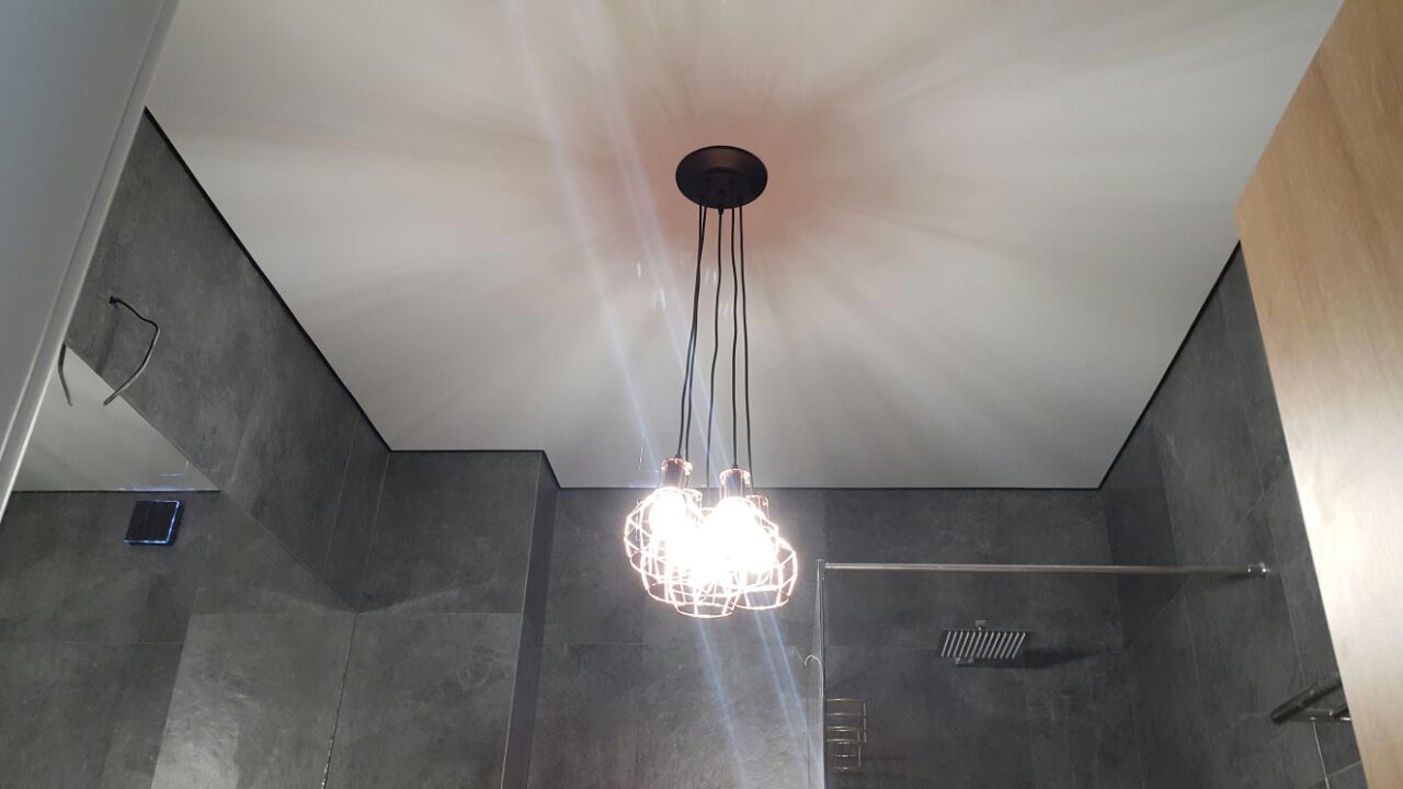 Biały sufit napinany w łazience z szarymi płytkami, oświetlony dekoracyjną lampą wiszącą z trzema żarówkami w metalowych kloszach.