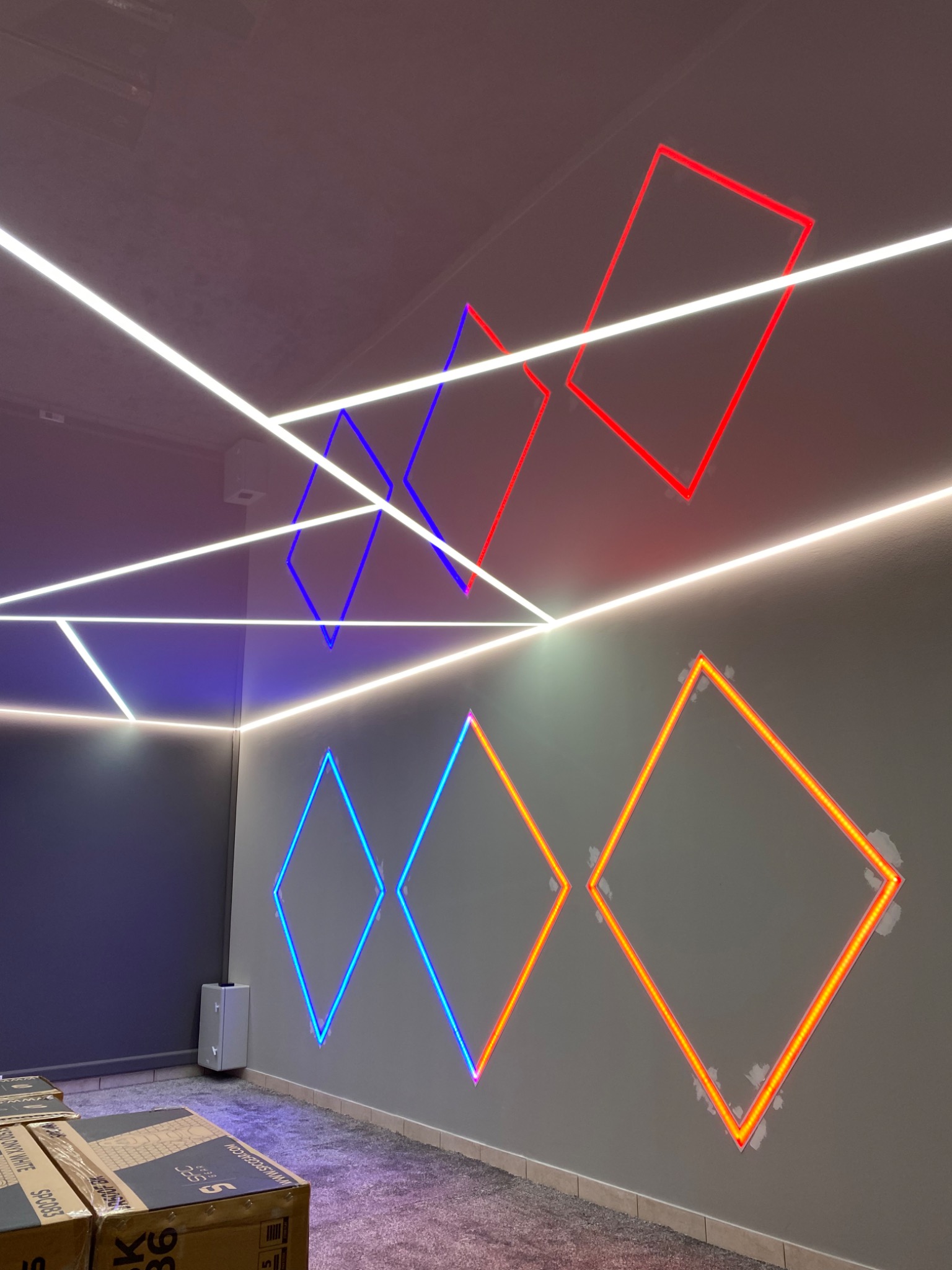 Nowoczesne oświetlenie sufitowe i ścienne z neonowymi kwadratami w kolorach czerwonym, niebieskim i pomarańczowym, widoczne ślady po montażu na ścianie.