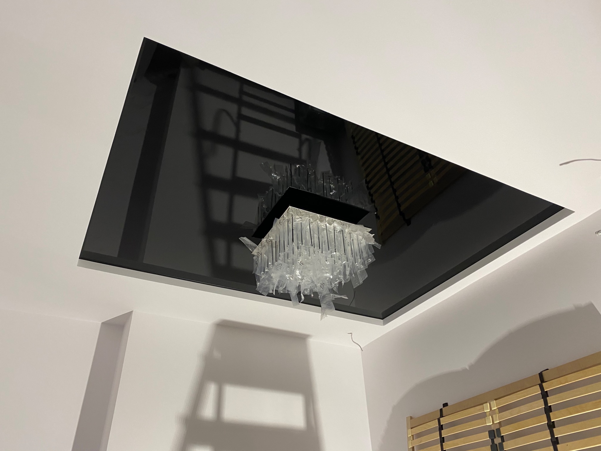 Błyszczący, czarny sufit napinany z wbudowanym oświetleniem w postaci żyrandola z elementami z tworzywa sztucznego jeszcze w folii ochronnej, widoczne fragmenty białych ścian i drewnianego zagłówka...