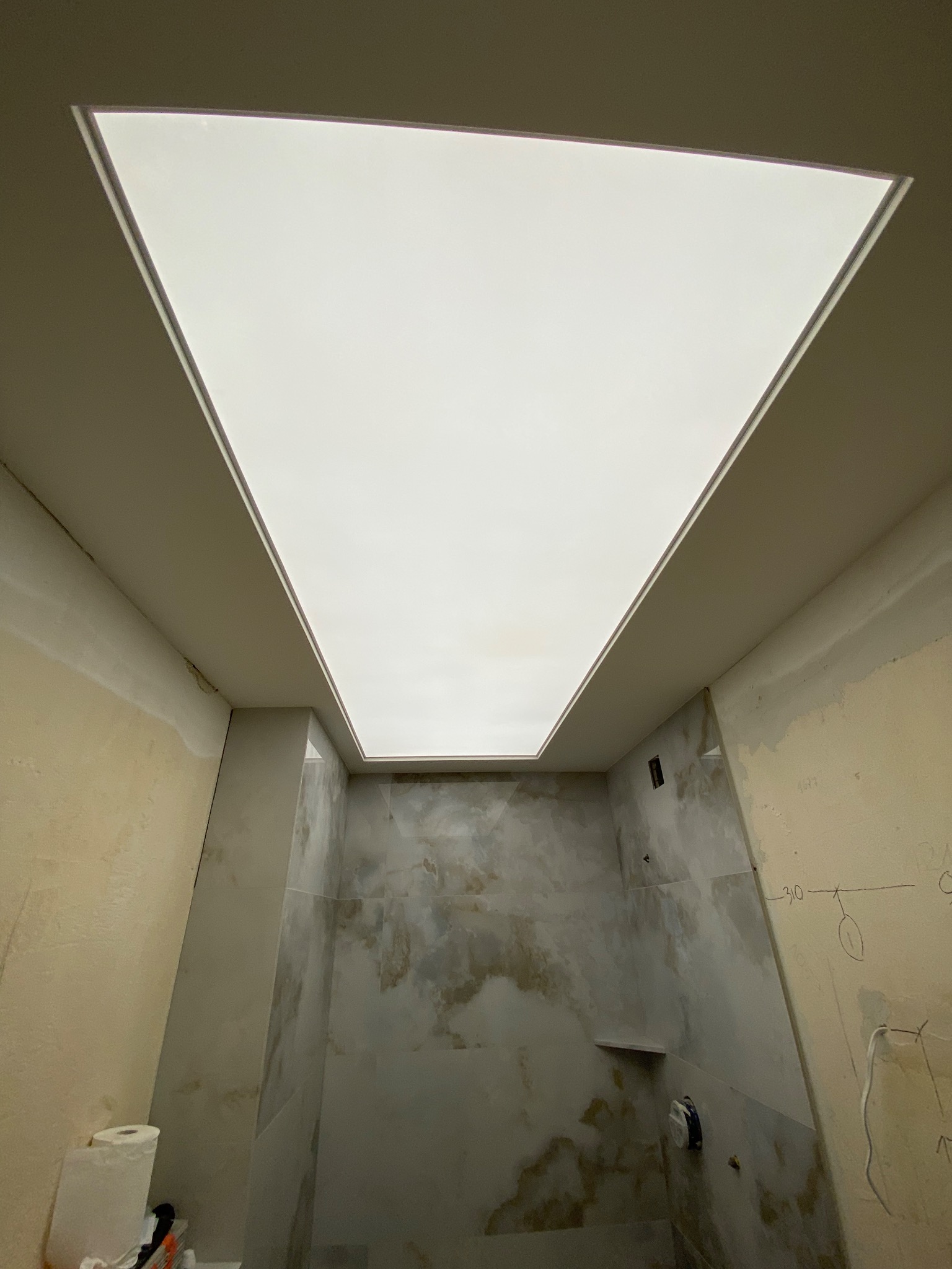 Narożne ujęcie łazienki w trakcie remontu, z zamontowanym podświetlanym sufitem napinanym w kształcie trapezu, widoczne surowe ściany z instalacjami i zapiskami, oraz rolka papieru i narzędzia w rogu.