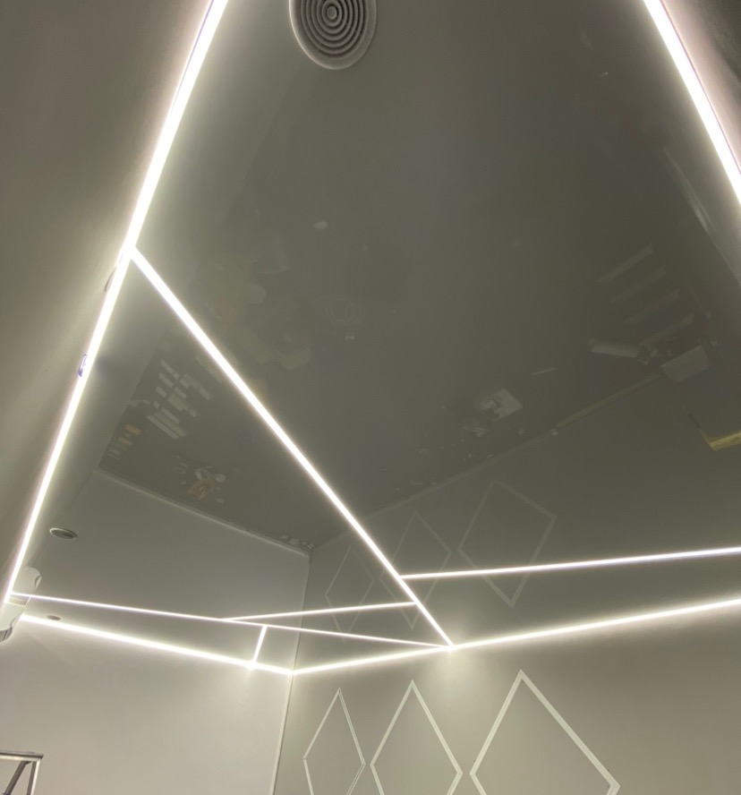 Nowoczesny sufit napinany z liniowym oświetleniem LED tworzącym geometryczne wzory na ścianach i suficie, widoczny okrągły nawiewnik wentylacyjny.