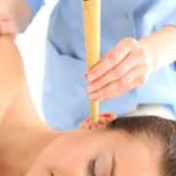 Kobieta leżąca na boku z zamkniętymi oczami podczas zabiegu świecowania uszu, trzymana przez osobę w niebieskim uniformie.