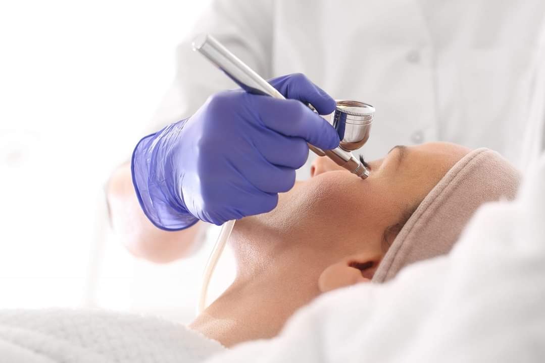 Osoba leżąca na leżance podczas zabiegu na twarz wykonywanego aerografem przez specjalistę w niebieskich rękawiczkach, w tle białe prześcieradło i fartuch.