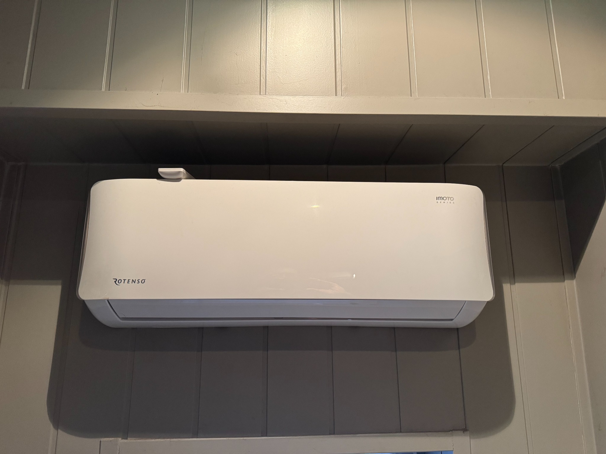 Biała klimatyzacja Rotenso Imoto zamontowana na szarej, panelowej ścianie w pomieszczeniu, widok z dołu, oświetlenie dzienne.