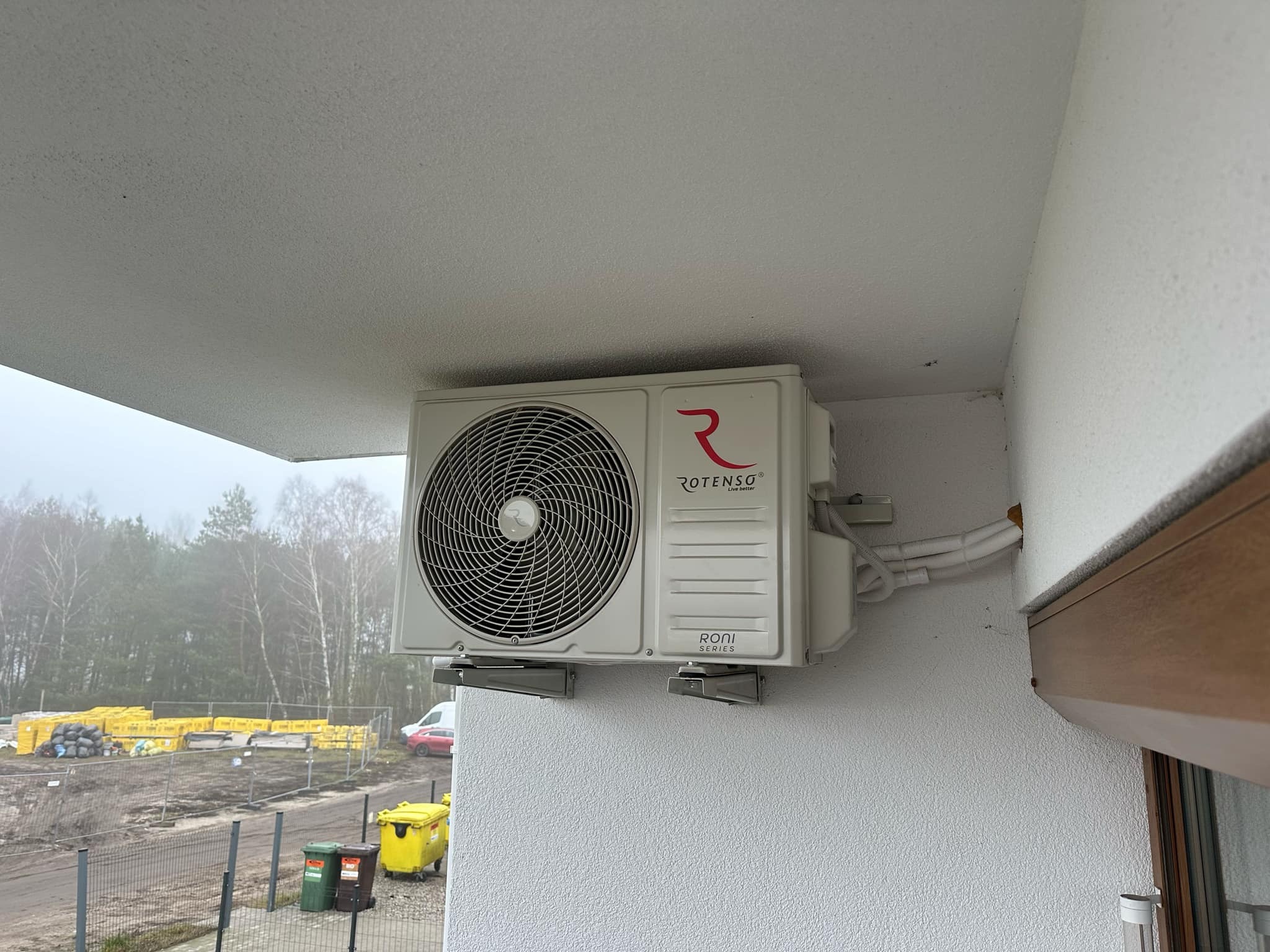 Zamontowana jednostka zewnętrzna klimatyzacji Rotenso Roni na elewacji budynku z widocznymi przewodami i otoczeniem placu budowy w tle.