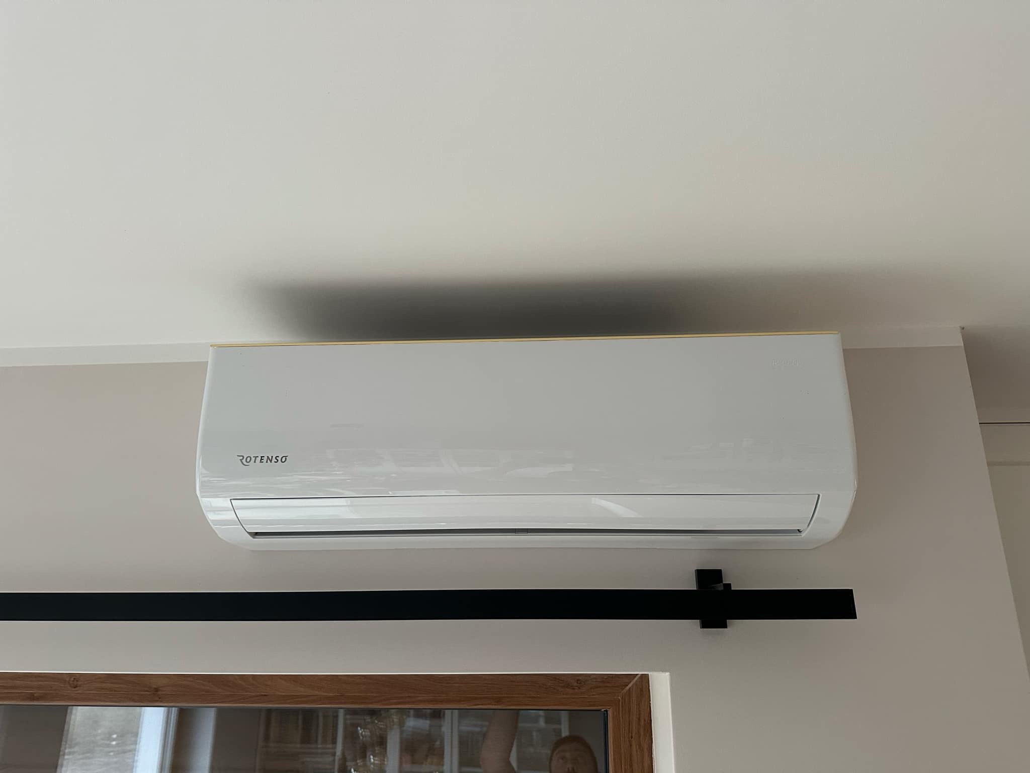 Nowoczesny, biały klimatyzator Rotenso zamontowany na ścianie w jasnym wnętrzu, widoczny fragment ramy okiennej i czarny element dekoracyjny.