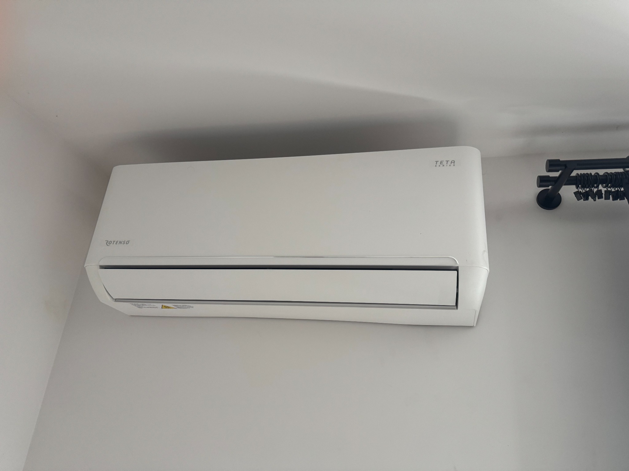 Biała klimatyzacja Rotenso Teta Series zamontowana na jasnej ścianie, widoczna z boku, z czarnym karniszem w tle po prawej stronie.