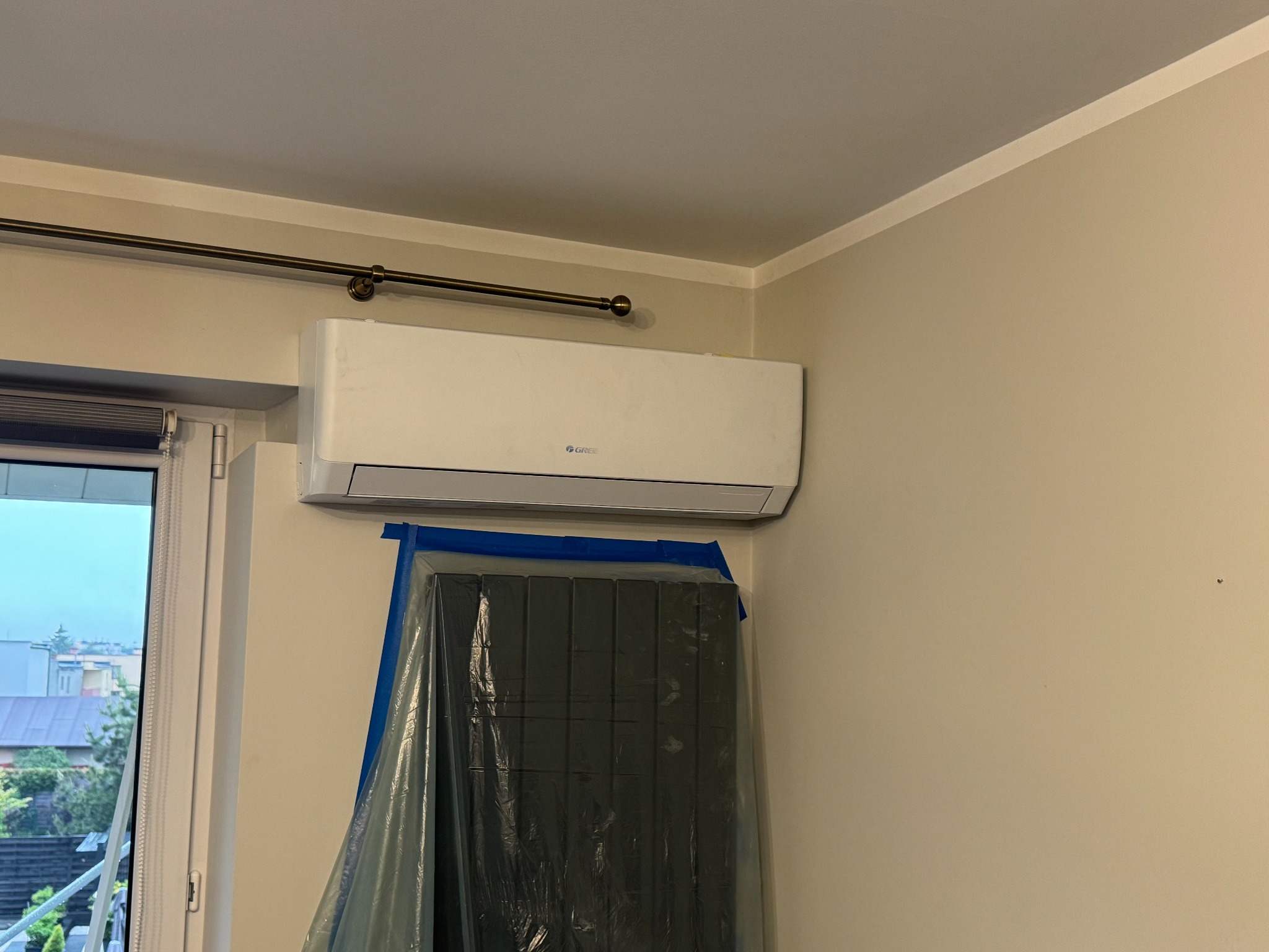 Biały klimatyzator marki Gree zamontowany na ścianie w pokoju, z zasłoniętym folią meblem poniżej. Widoczny fragment okna i karnisz.