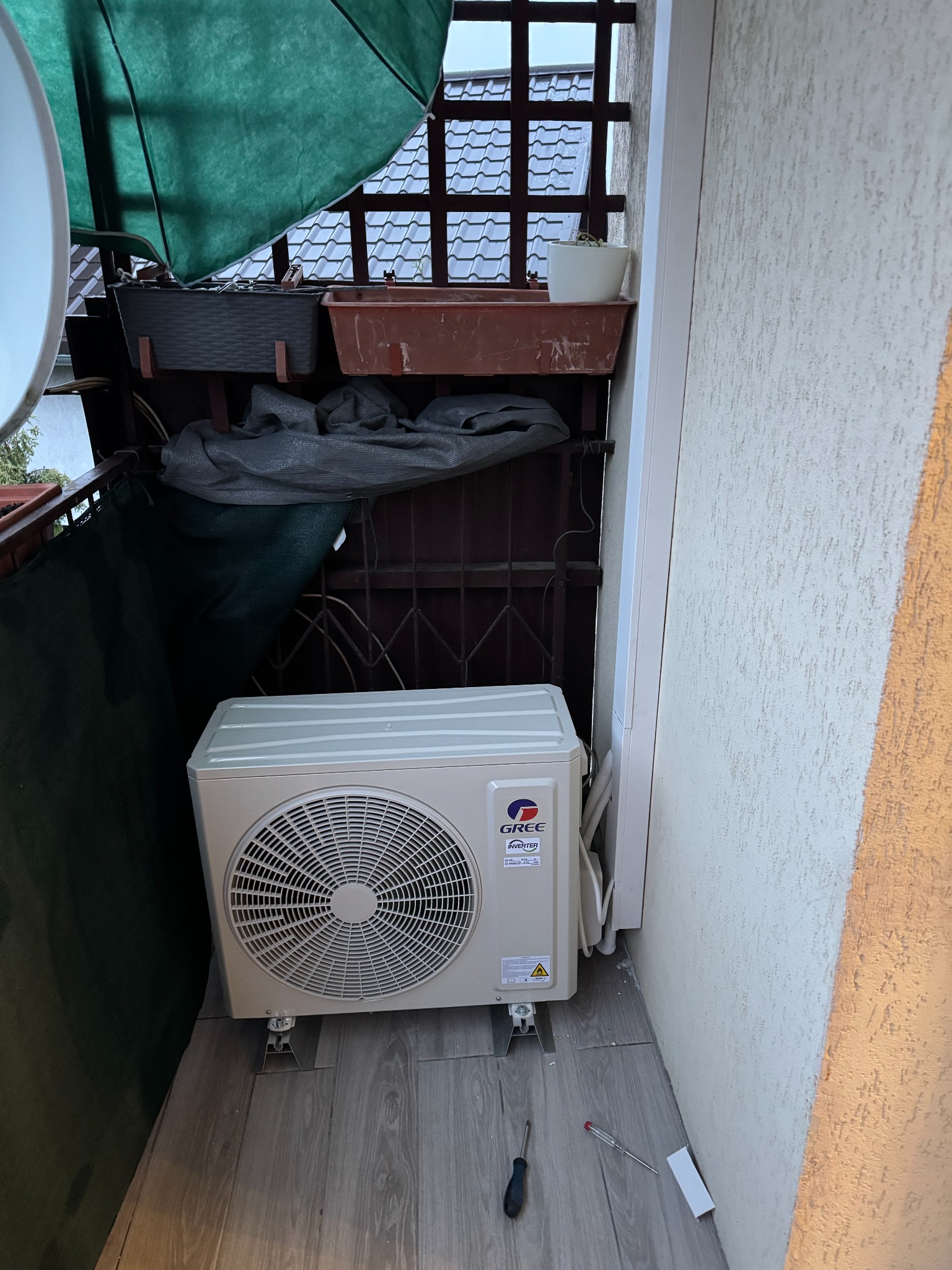 Zamontowana jednostka zewnętrzna klimatyzacji GREE na balkonie, widoczne narzędzia. Na górze balkonowe donice i trejaż.