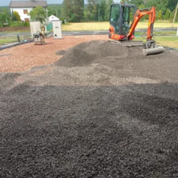 Pomarańczowa mini koparka Kubota na stercie ciemnego gruzu, w tle czerwony gruz, zagęszczarka i domy jednorodzinne.